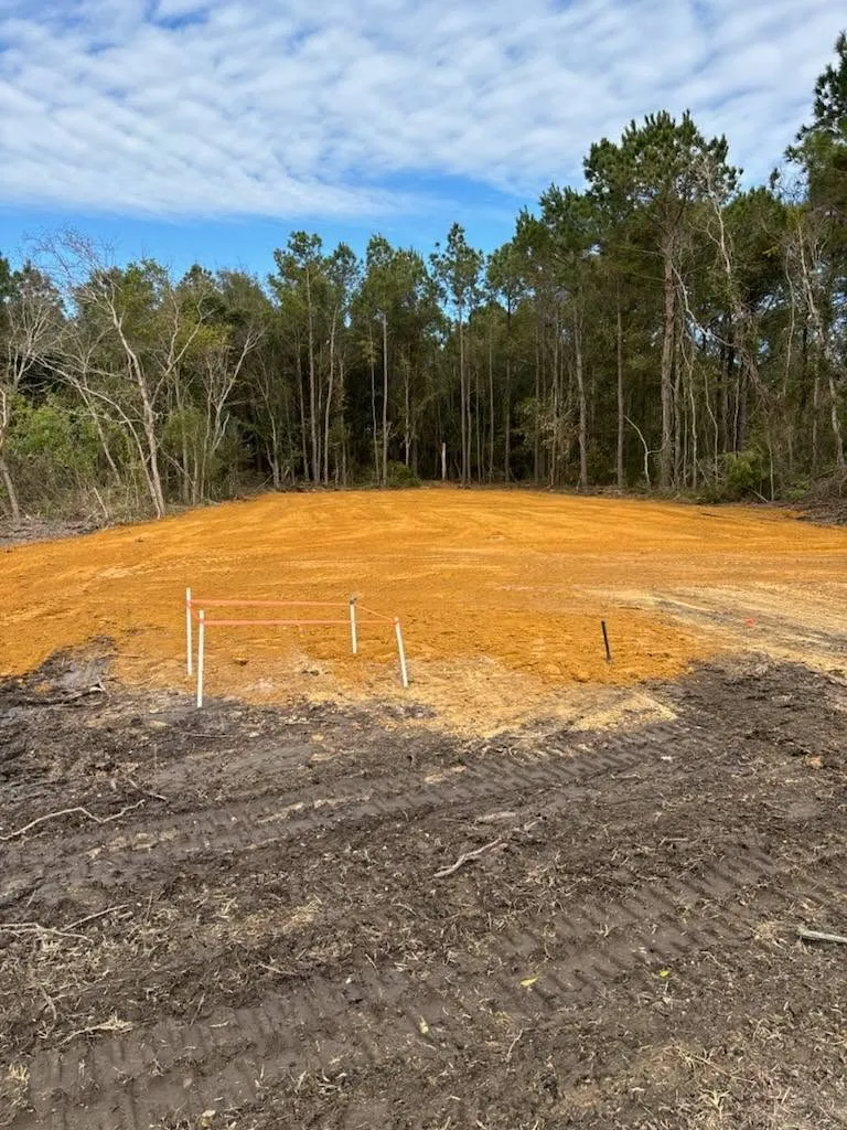 Prepared orange-earth pad bordered by woodland with layout stakes