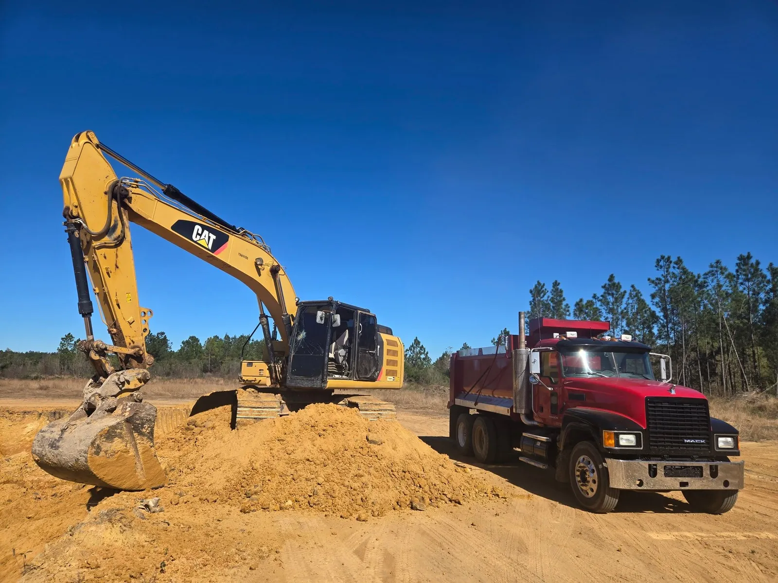 CAT excavator and red dump truck beside a pile of excavated earth under blue sky
